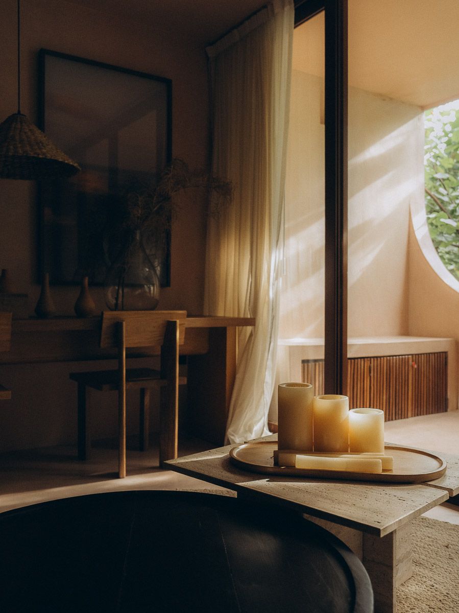A dark freestanding tub beside a travertine table holding a tray of pillar candles, afternoon light through a round window