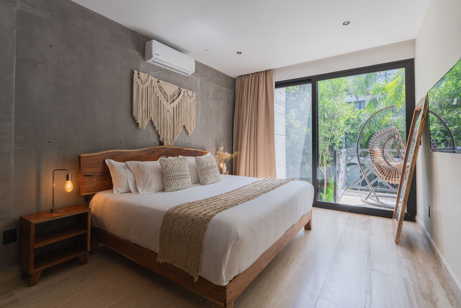 A bedroom with a live-edge wood headboard and concrete accent wall, open sliding doors onto a balcony where a round hanging rattan chair sits among palms and greenery