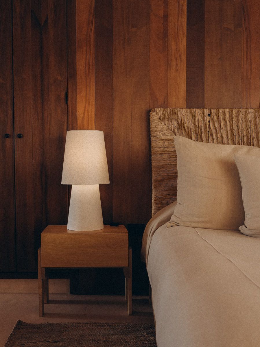 A lit ceramic table lamp on a wood nightstand against a walnut-panelled wall, beside a bed with a woven headboard