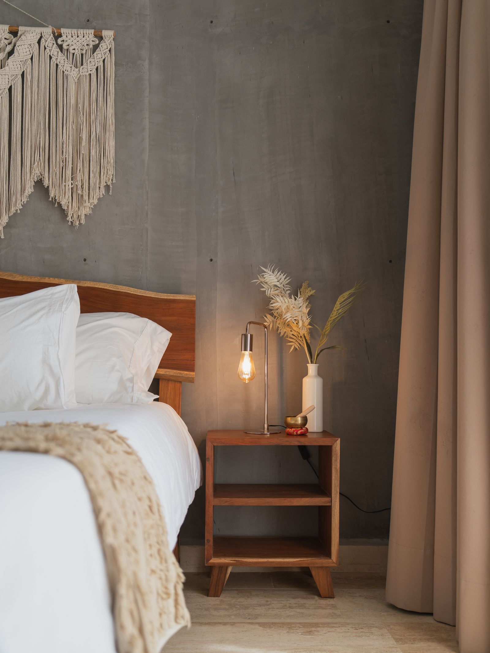A lit brass task lamp on a wood nightstand beside a bed, with a macramé wall hanging above, a vase of dried pampas grass, and a small brass singing bowl
