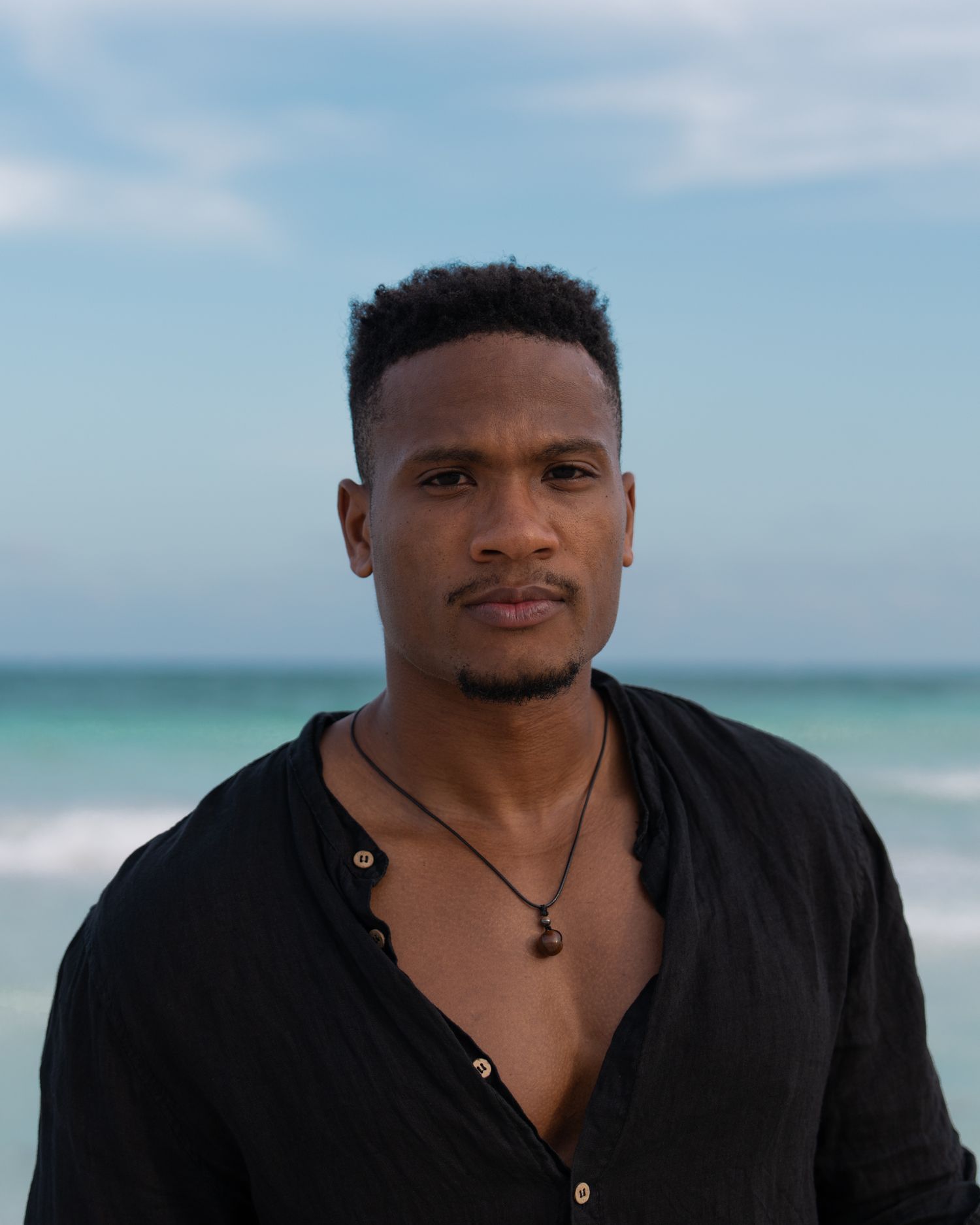 Man in an open black linen shirt standing before turquoise water, direct gaze