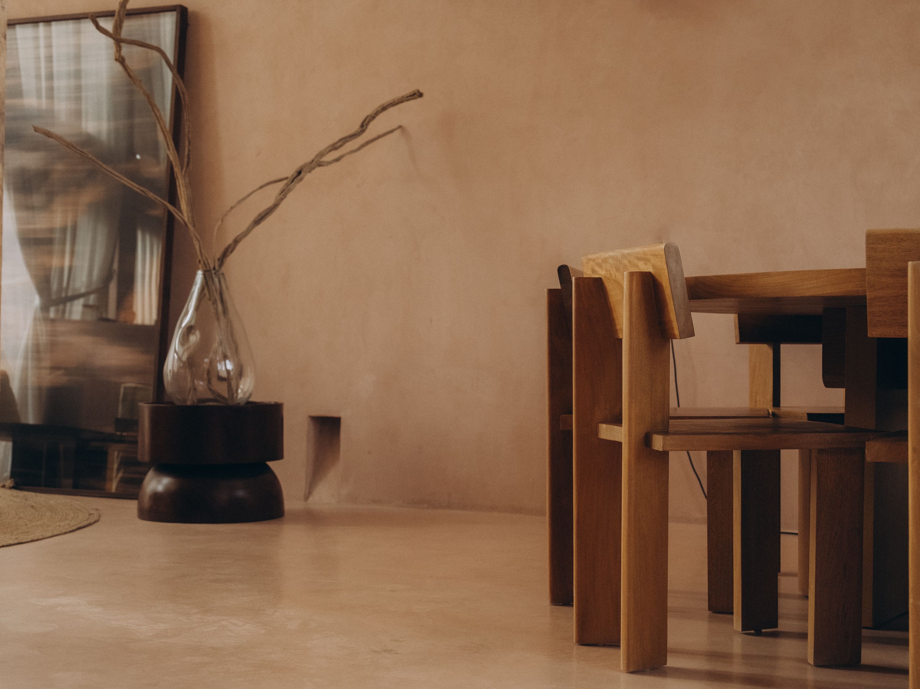 Carved wood dining chairs against a plaster wall, a glass vase of bare branches beside a leaning mirror