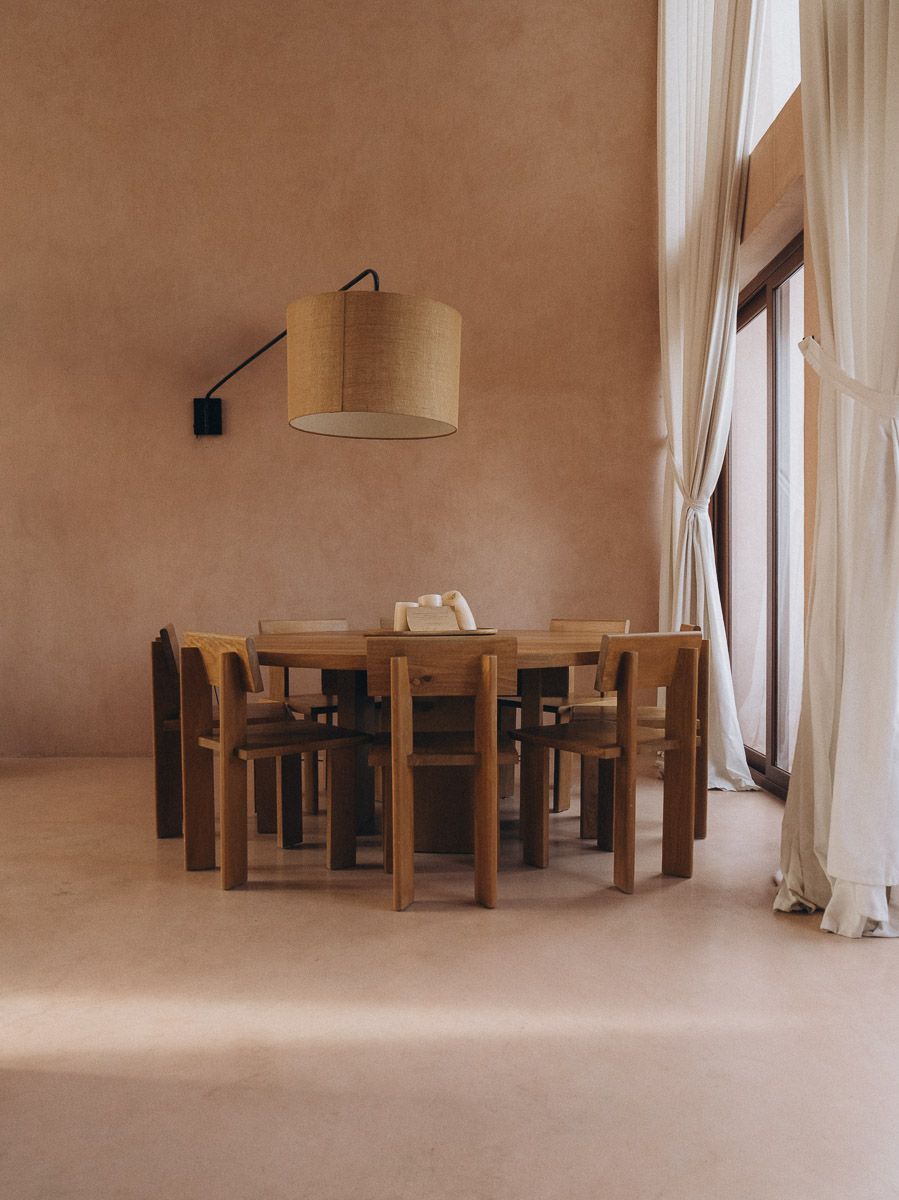 A round wood dining table surrounded by carved chairs beneath a wall-mounted drum-shade lamp against a terracotta wall