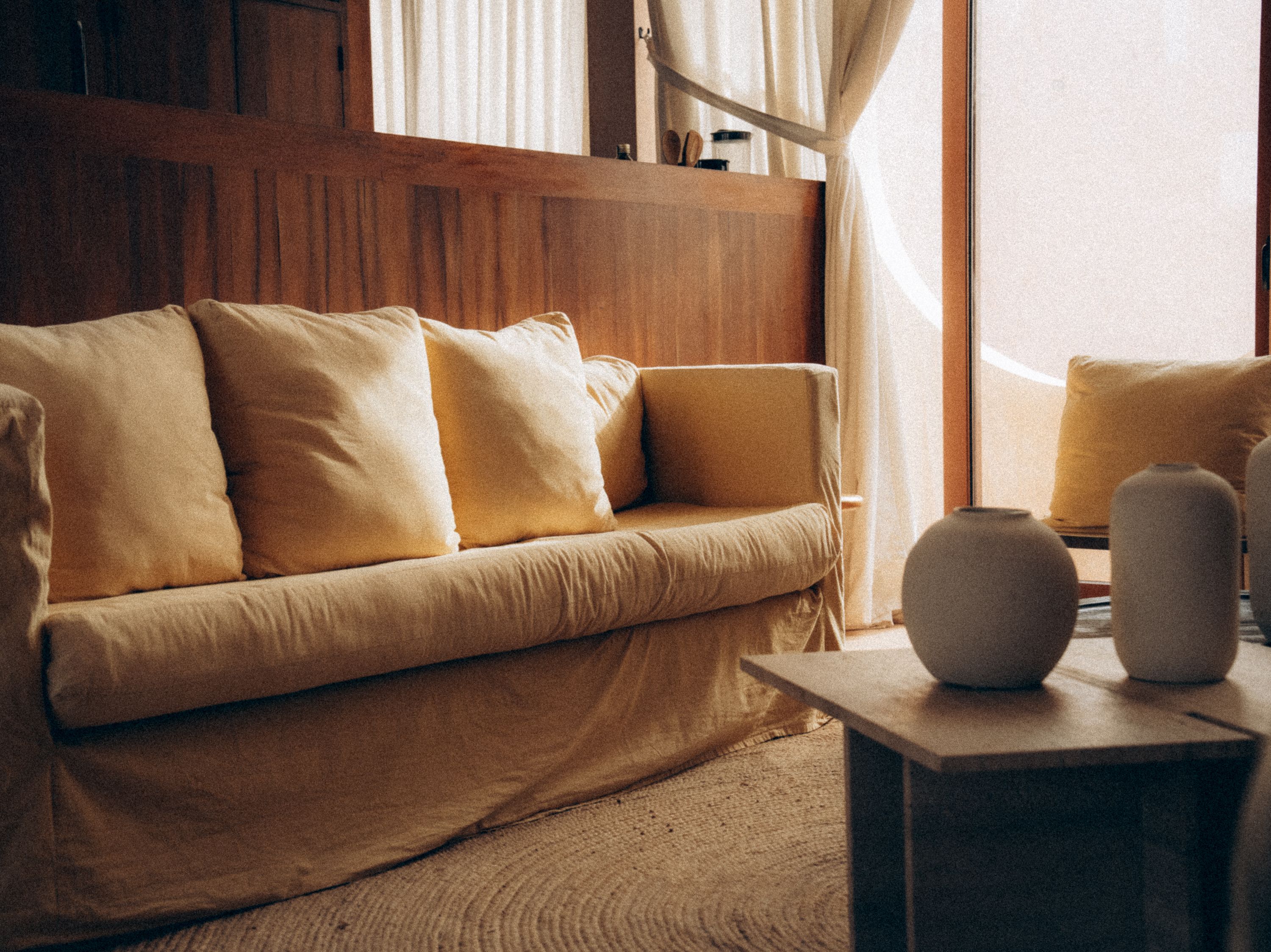 A linen-slipcovered sofa with butter-yellow pillows against a wood-panelled wall, ceramic vases on a low table beside sheer curtains