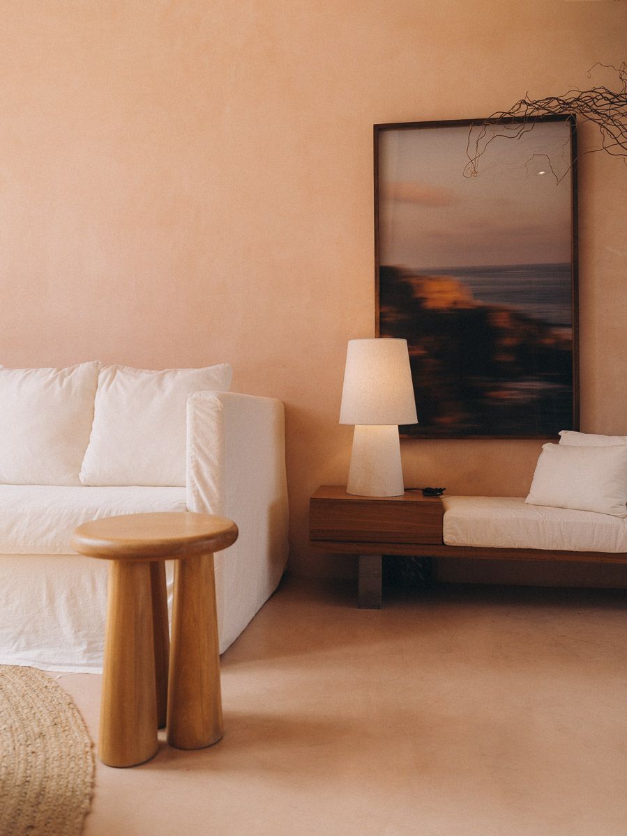 A white linen sofa and turned-wood stool beside a walnut bench, a framed coastal photograph lit by a ceramic table lamp