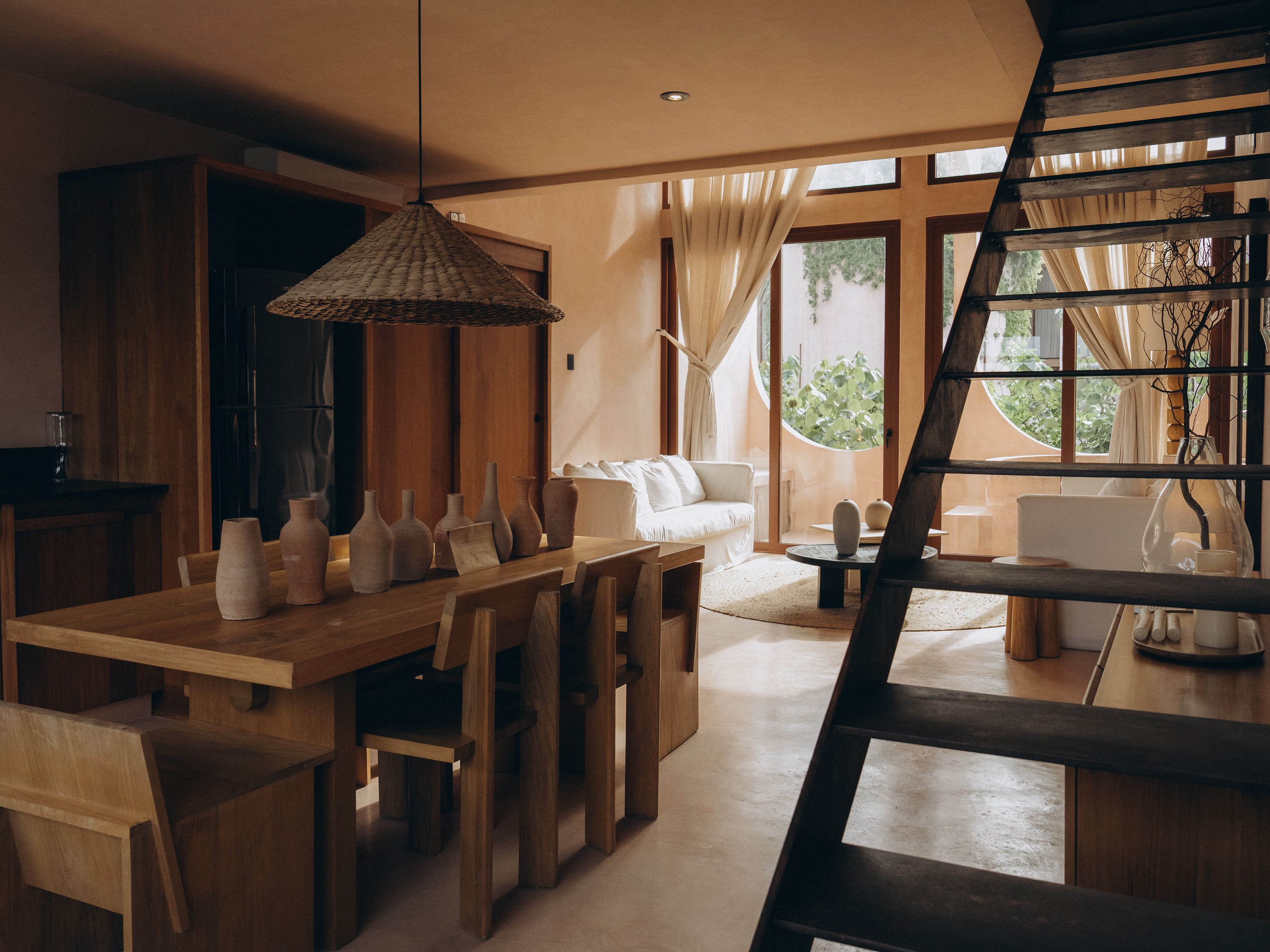 A terracotta-toned dining room with a woven pendant lamp over a wood table set with ceramic vases, a dark timber staircase rising beside it