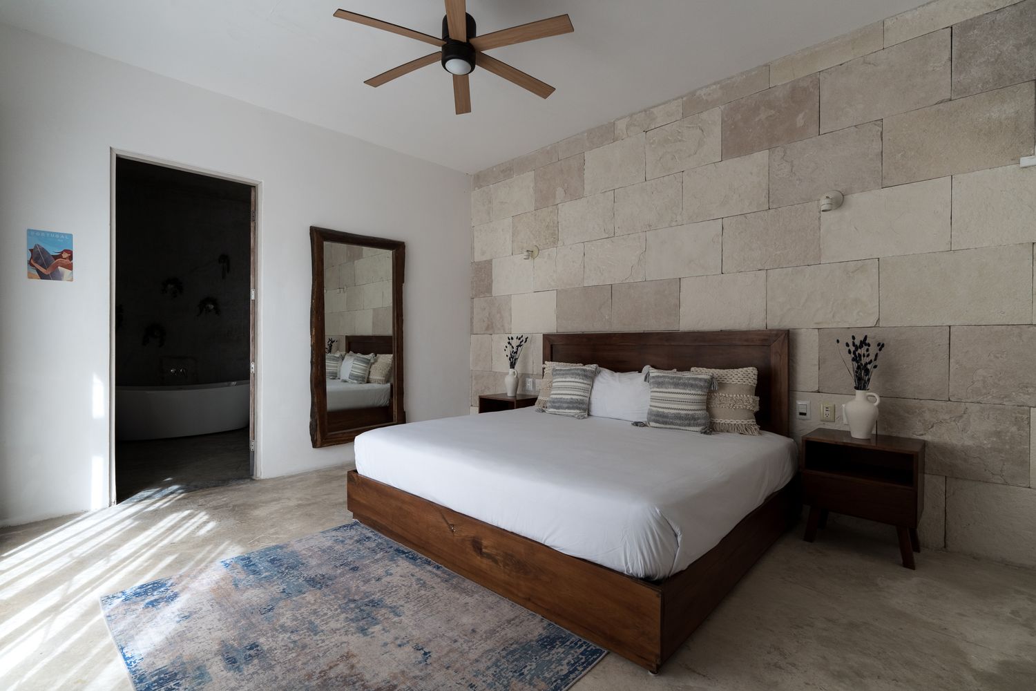 Bedroom with a rough limestone block accent wall behind the bed, a wood ceiling fan, a tall leaning floor mirror, and a doorway open to an ensuite bathroom with a freestanding tub