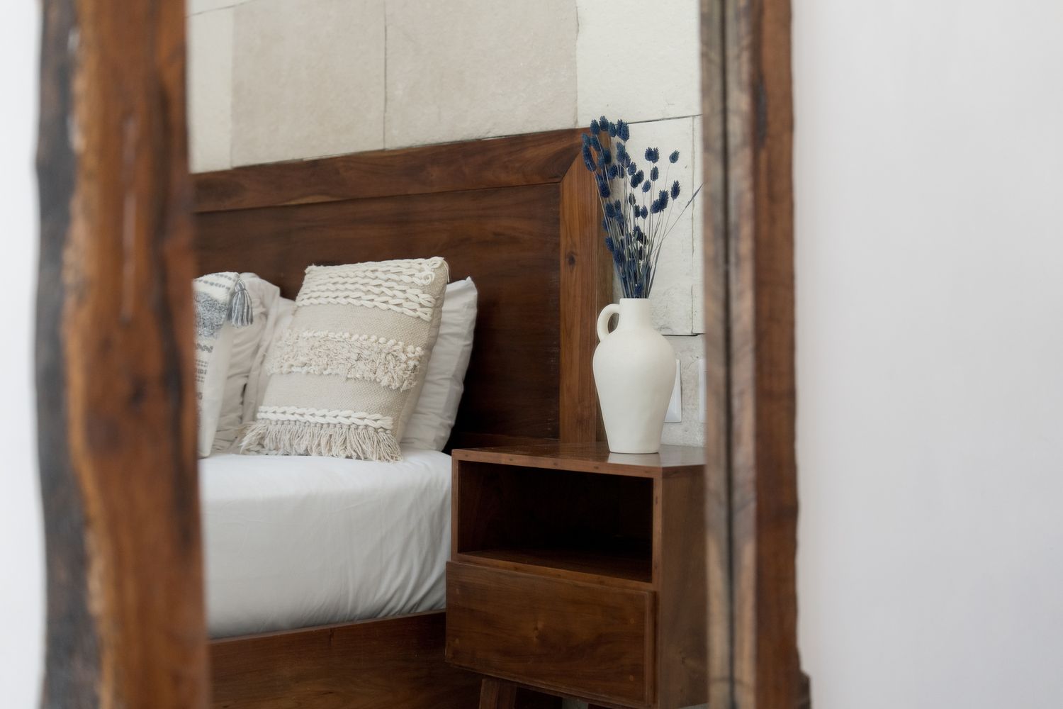 A tall wood-framed floor mirror reflecting a bed with white linens and a fringed woven pillow, a walnut nightstand, and a white ceramic jug holding dried lavender