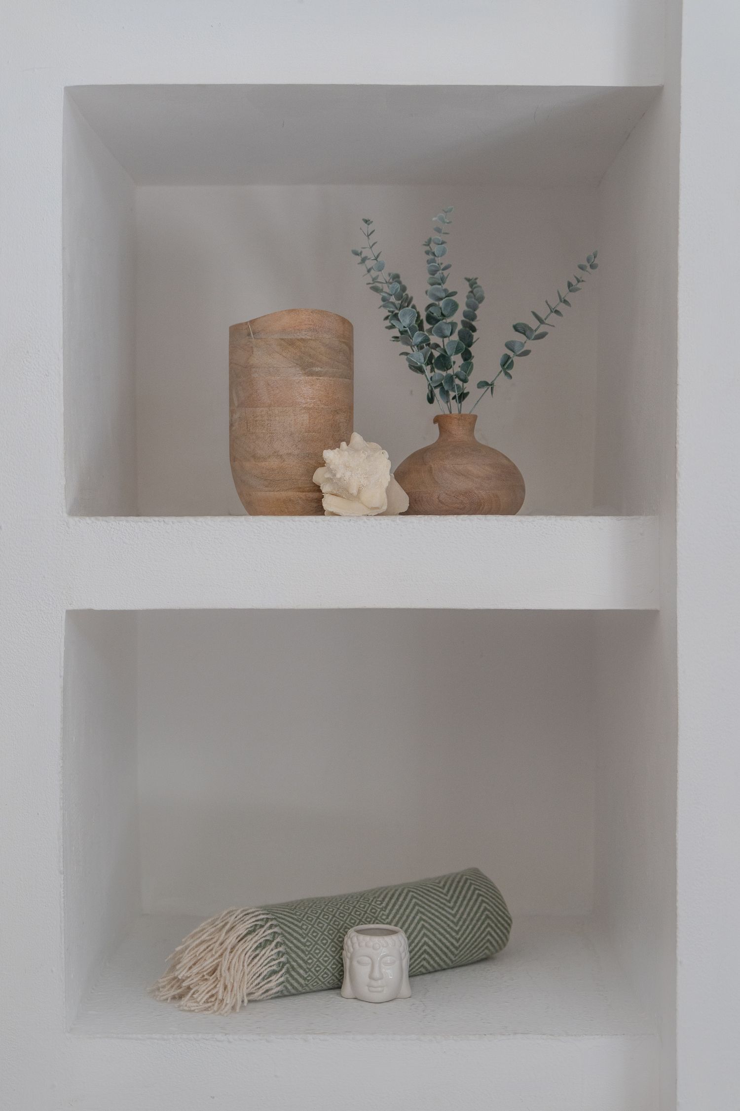 Two built-in wall niches: the upper shelf holds a turned wood vase, a bud vase of dried eucalyptus, and a piece of white coral; the lower shelf holds a rolled green herringbone throw and a small ceramic buddha-head candle holder