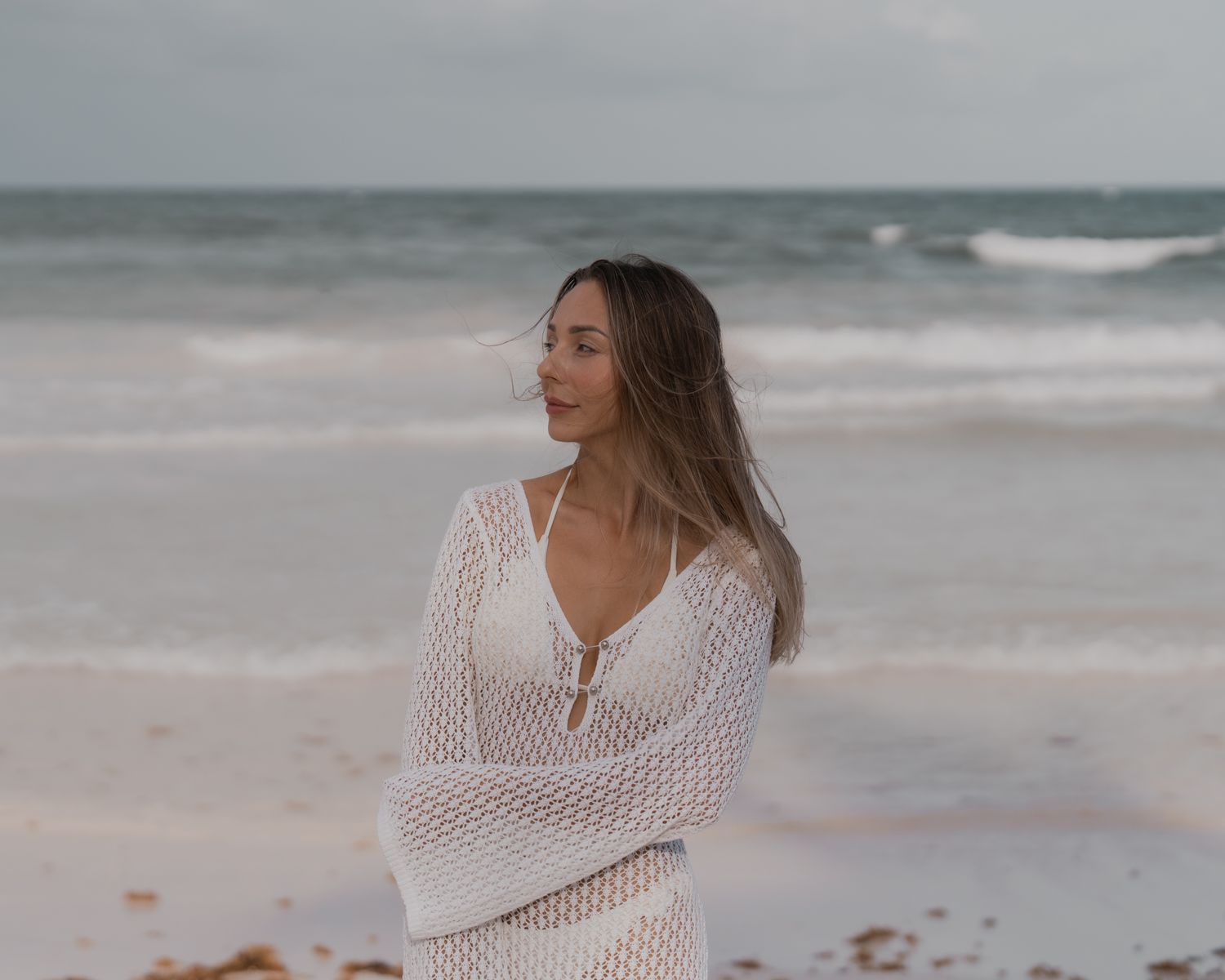 Woman in a white crochet cover-up on an overcast beach, hair blowing in the wind, looking off to the side